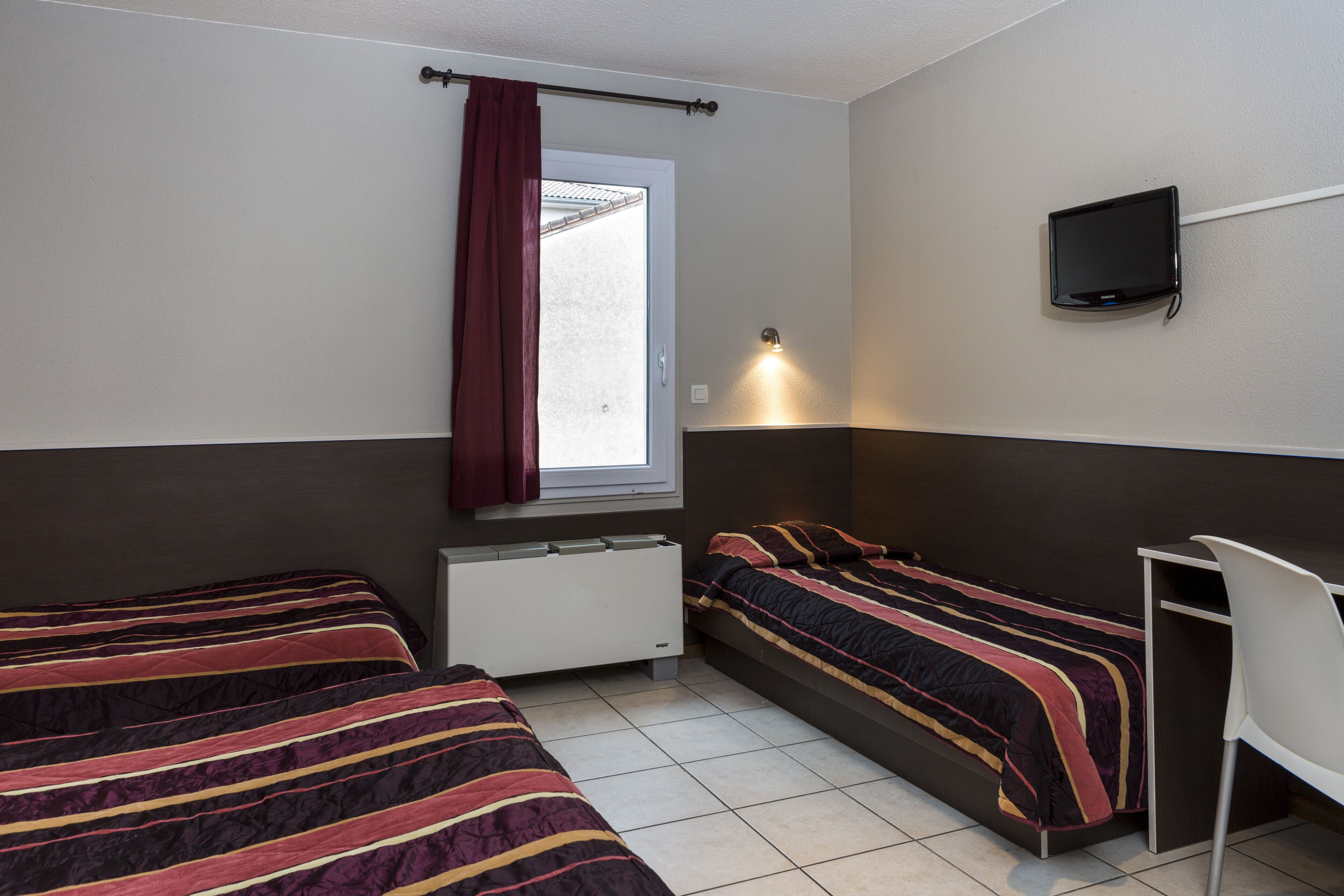 A small, well-lit room with two beds, a desk, a TV, and a window with a curtain.