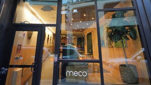 Entrada de un hotel con fachada moderna de vidrio, interior visible con mostrador de recepción y un letrero que indica el nombre 'meco hotel'.