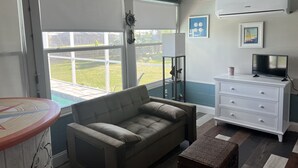 A living room with a sofa, a television, a white dresser, and a window overlooking a garden. - Waterfront Sea Cottage with private Pool and waterfront views (Port Charlotte)