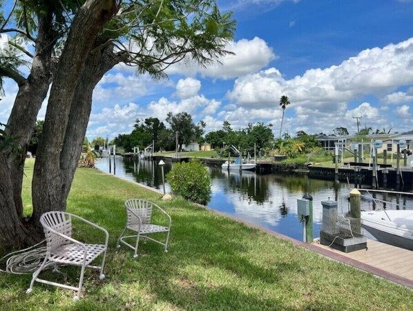 A riverside view with a tree, two metal chairs, and a dock with boats.