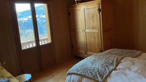 A bedroom with a wooden wardrobe, a bed with a white blanket, a small table, and a window with a view of mountains. - chalet with breathtaking views of the alps (Crans-Montana)