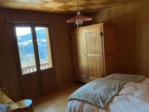 Un dormitorio con un armario de madera, una cama con manta blanca, una mesita y una ventana con vistas a las montañas.