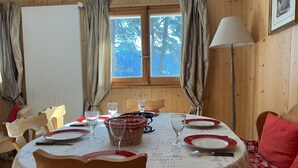 A dining area with a table set for four, wooden chairs, a window with curtains, and a lamp. - chalet with breathtaking views of the alps (Crans-Montana)