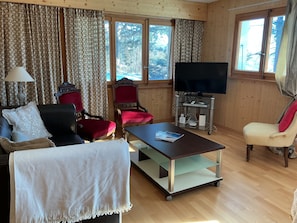 Una sala de estar con sofá, sillones, una mesa de centro, televisor en un mueble bajo y una ventana con cortinas.