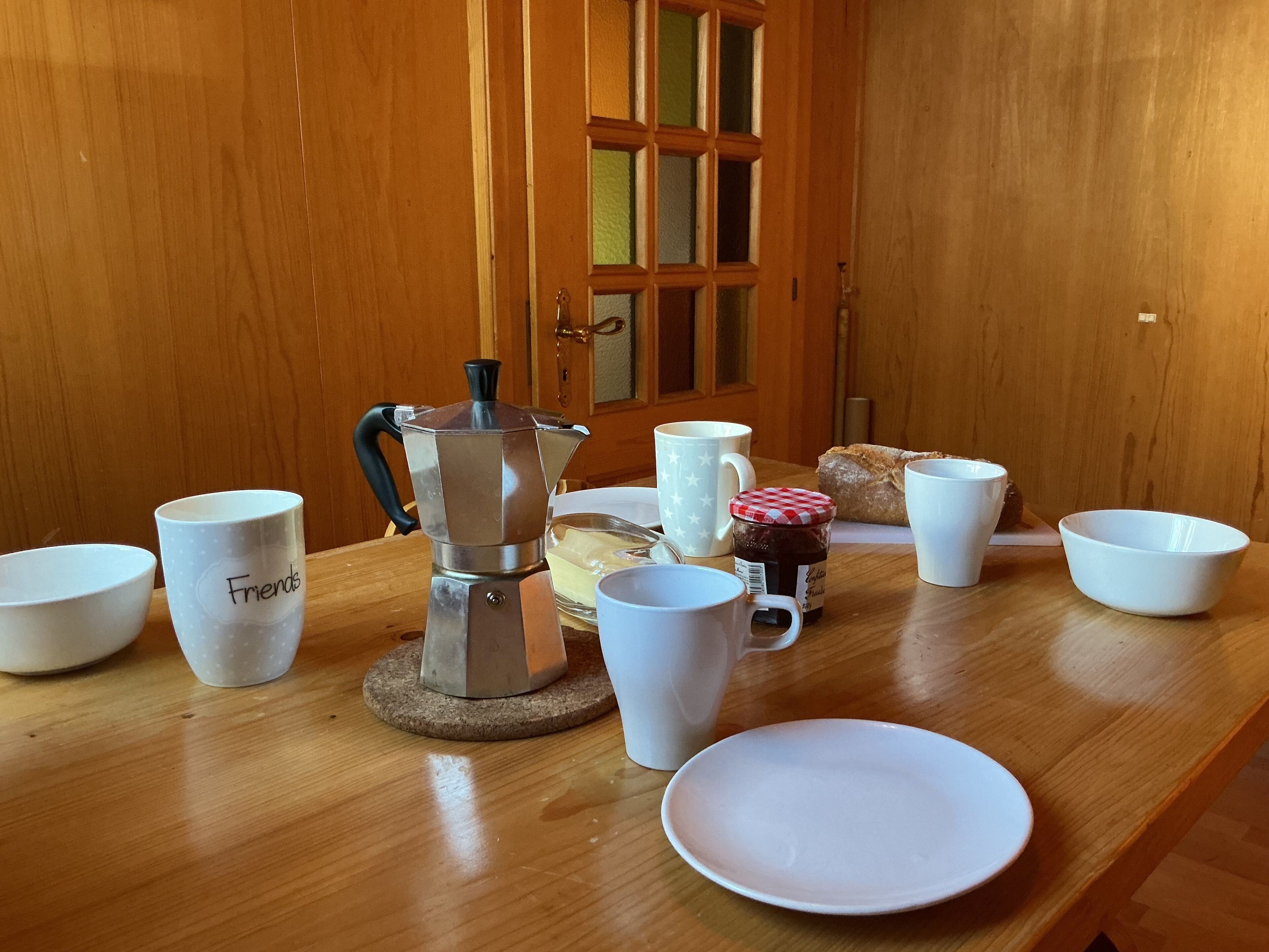 Ein Holztisch mit Kaffeemaschine, Tassen, einem Teller und einem Marmeladenglas.