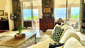 Una sala de estar con una pared de piedra, un sofá blanco con cojines estampados, un mueble de madera y vistas al océano a través de puertas correderas grandes.