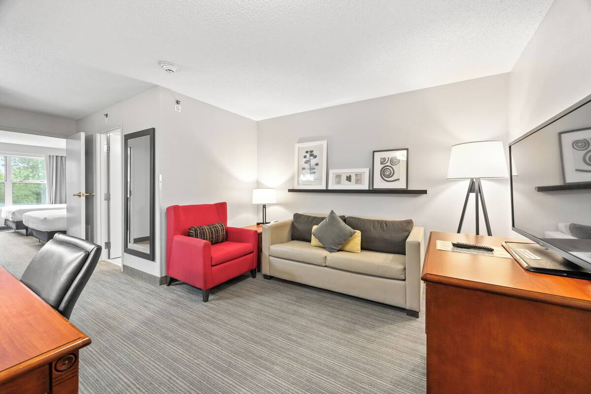 A hotel room with a wooden desk, a red armchair, a beige sofa, and a TV on a wooden stand.