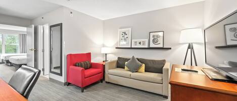 Habitación de hotel con un escritorio de madera, un sillón rojo, un sofá beige y un televisor sobre un mueble de madera.
