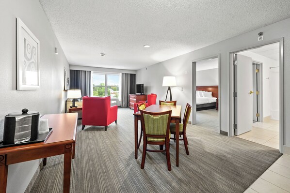 A hotel room with a bed, a desk with a lamp, a dining area with chairs, and a view of the outdoors. - Independence Stay Hotel & Suites (Marinette)