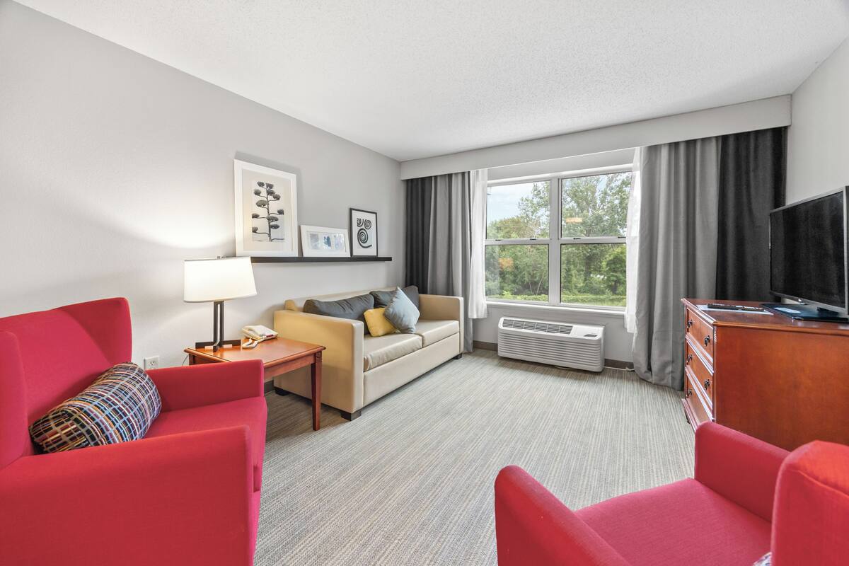 A hotel room with a red sofa, a beige sofa, a TV, a lamp, a small table, and a window with curtains.