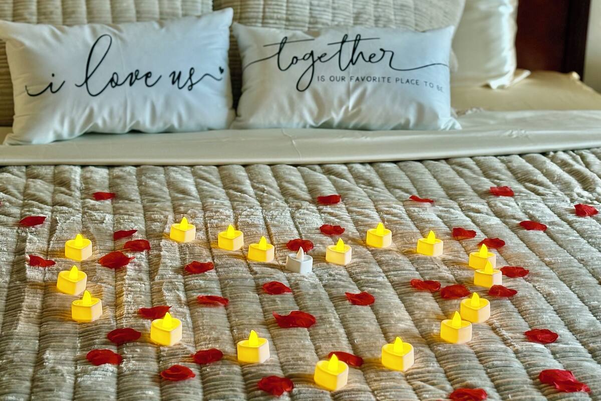 A bed with white bedding, two pillows with text, and a quilt with yellow candles and red rose petals.