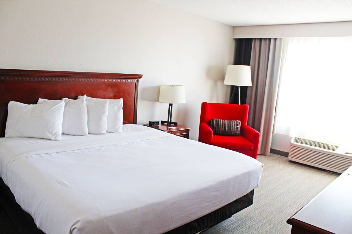 A hotel room with a large bed, a red armchair, a nightstand with a lamp, and an air conditioning unit.