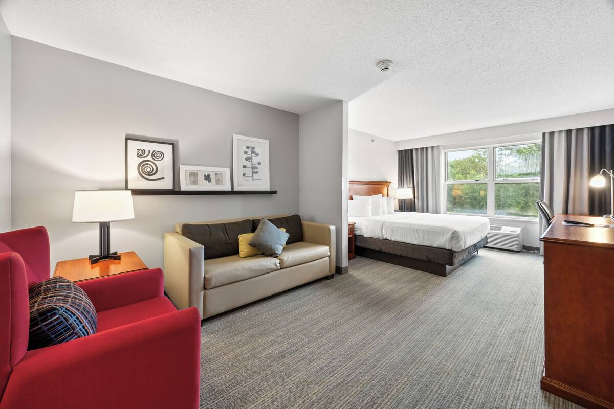 A hotel room with a bed, a sofa, a red armchair, a desk, and a window with curtains.