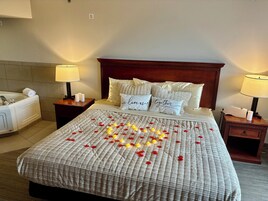 A hotel room with a bed, two bedside tables, a nightstand, a lamp, a towel, and a bath tub.