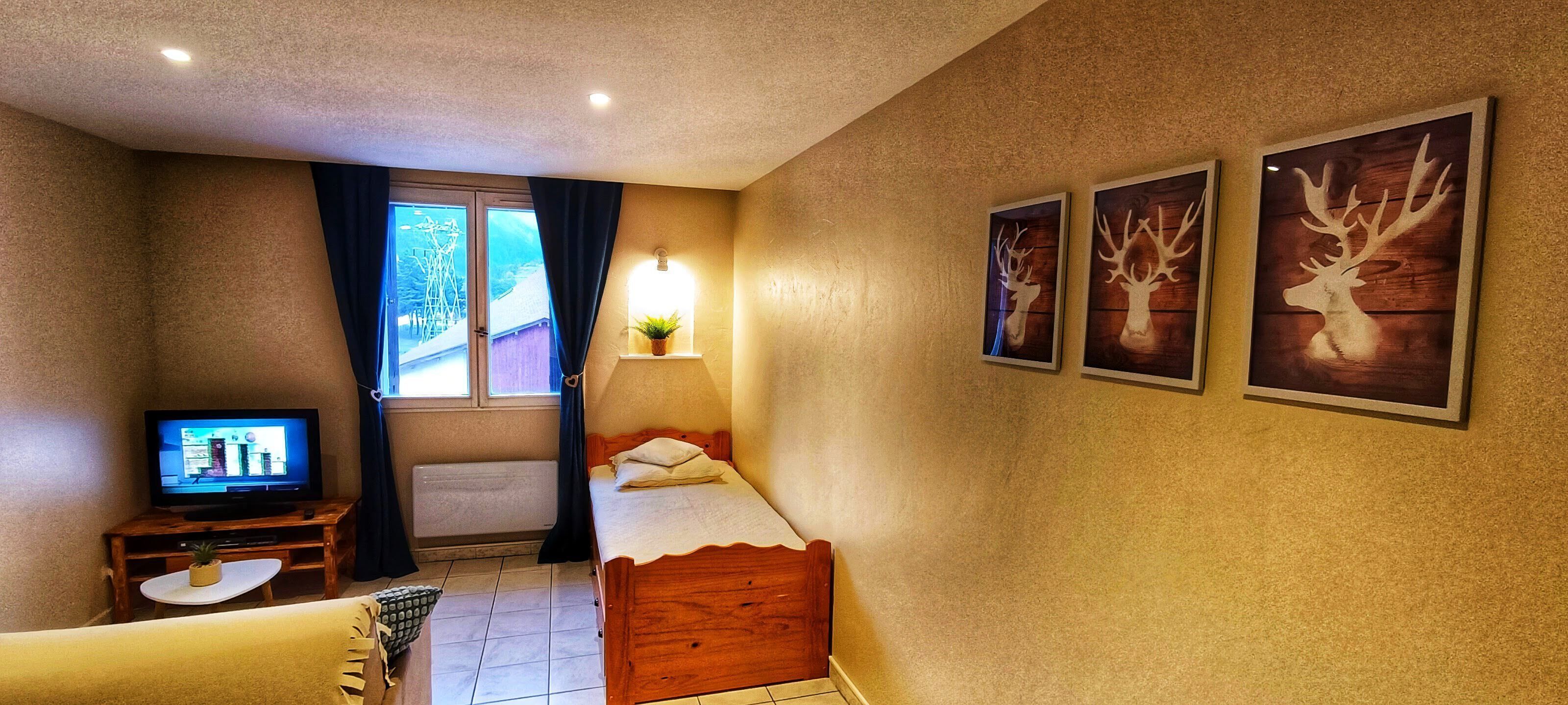 A small, cozy room with a bed, a TV, a window with curtains, and wall-mounted artwork.