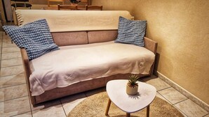 A beige sofa with patterned pillows, a small white table with a plant, and a tiled floor. - Grand Studio 3 Personnes Idéalement Placé (Barcelonnette)