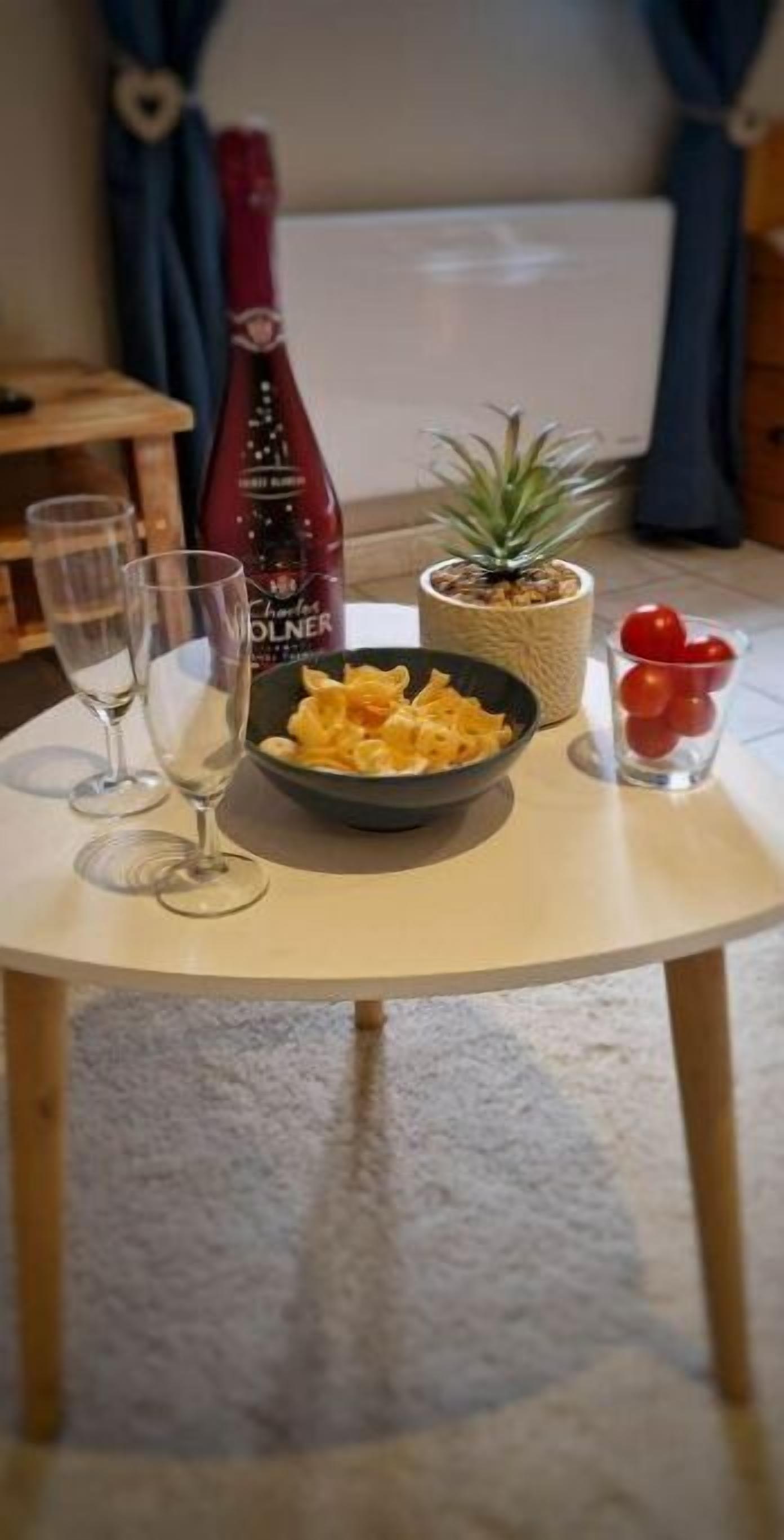 A small round table with a bowl of chips, a bottle of wine, a potted plant, and a glass of red grapes.