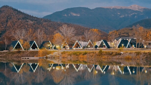 Cabañas con techo triangular a orillas de un lago, con reflejos, rodeadas de árboles otoñales y montañas al fondo.