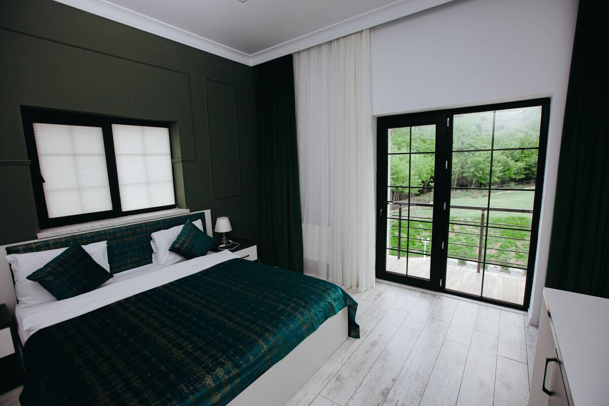 A bedroom with a large bed, a nightstand, a window with blinds, a sliding glass door to a balcony, and a view of greenery outside.