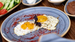 Una mesa de comedor con varios platos, incluyendo un plato con dos huevos fritos, tazones de salsas blanca y marrón, y un bol con pepinos y tomates en rodajas.