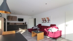 Una sala de estar moderna con un sofá rojo, una mesa de centro de madera y una mesa de comedor. Se observa una zona de cocina con armarios oscuros y un televisor de pantalla plana montado sobre la encimera.