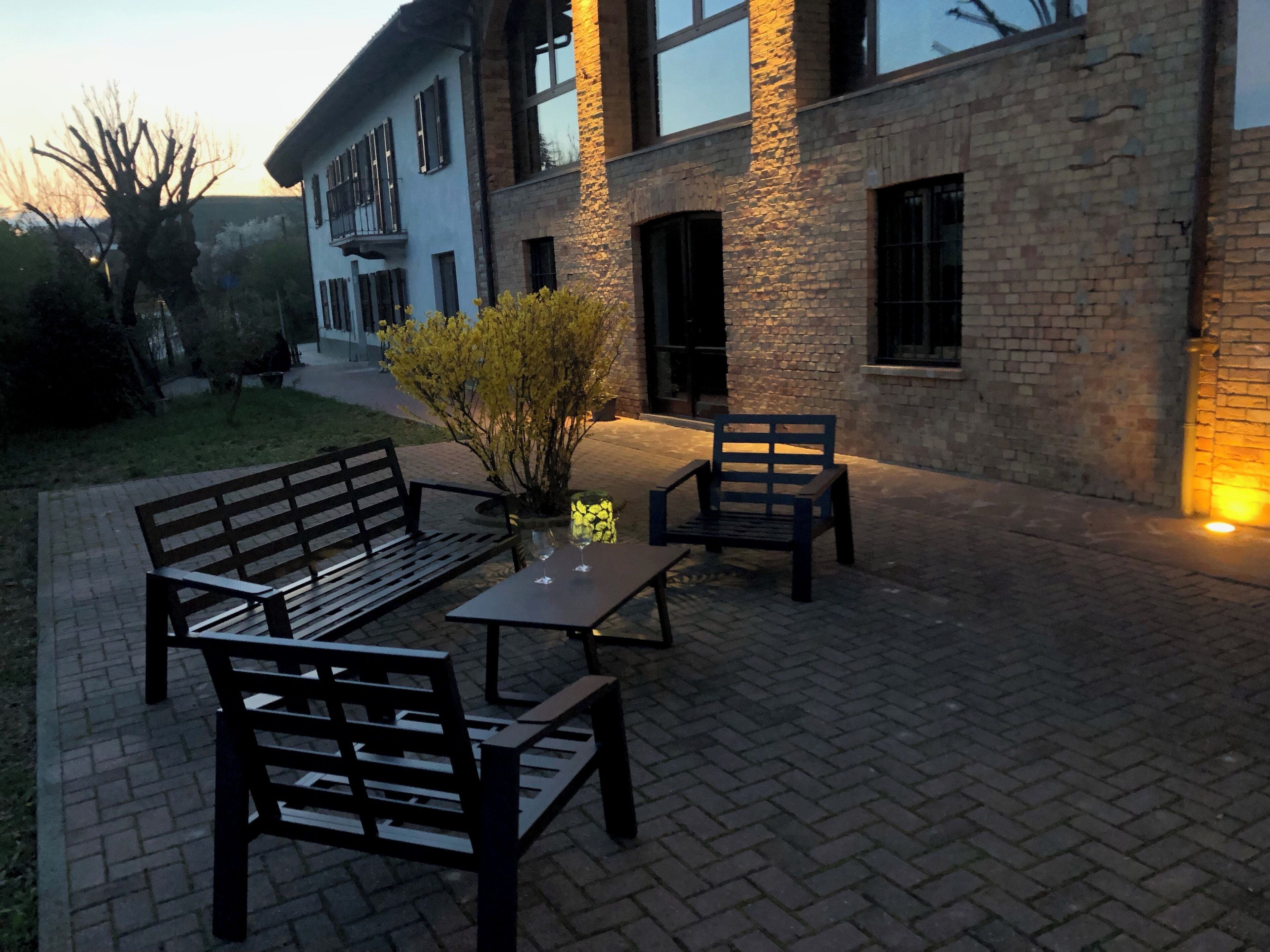 Un patio al aire libre con suelo de ladrillo, dos bancos de madera, una mesa con dos copas de vino y una planta en maceta.