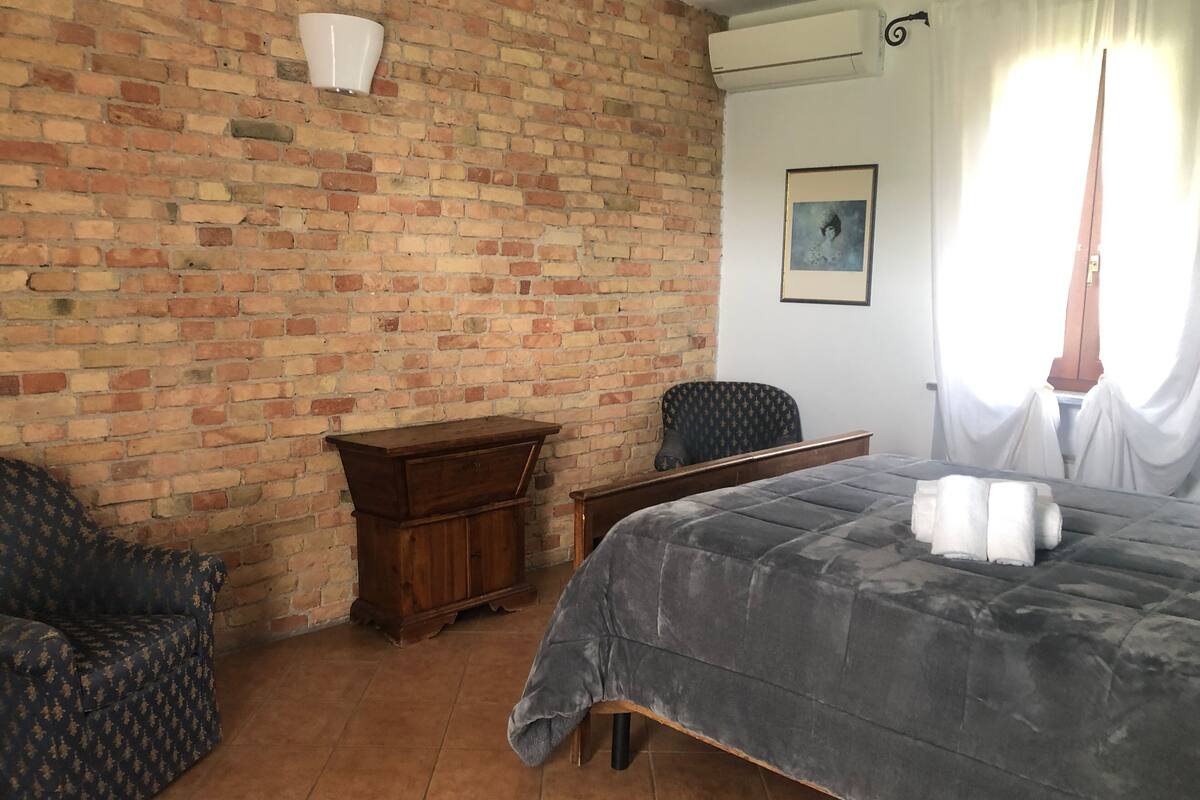 A room with a brick wall, a bed with a grey duvet, a wooden cabinet, a patterned armchair, and a chair.