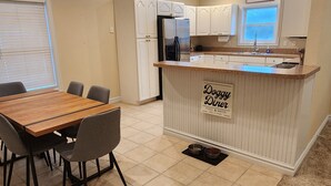 A modern kitchen with a central island labeled "Dogu Dining" and a dining area with a wooden table and chairs. - Goldie St Retreat - Heart of KDH, hot tub, pet friendly, centrally located  (Kill Devil Hills)