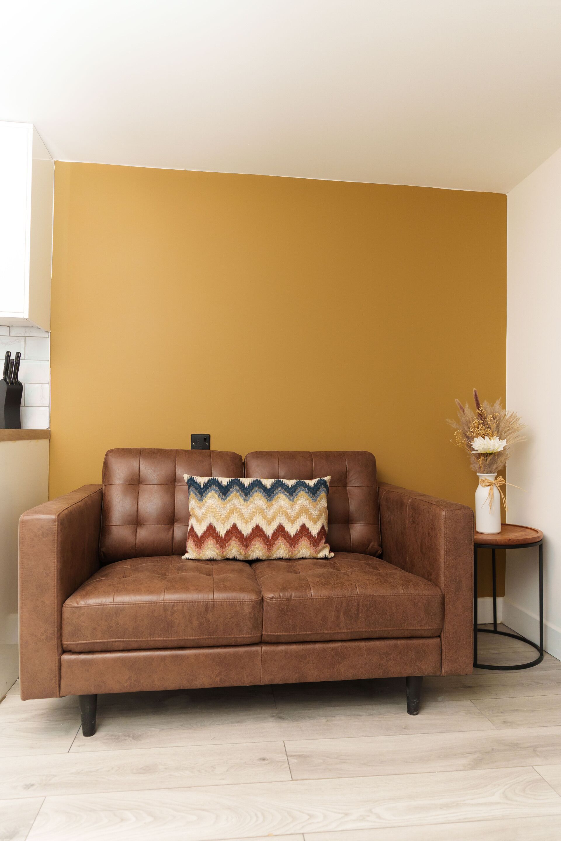 A brown leather sofa with a zigzag patterned pillow against a mustard yellow wall.