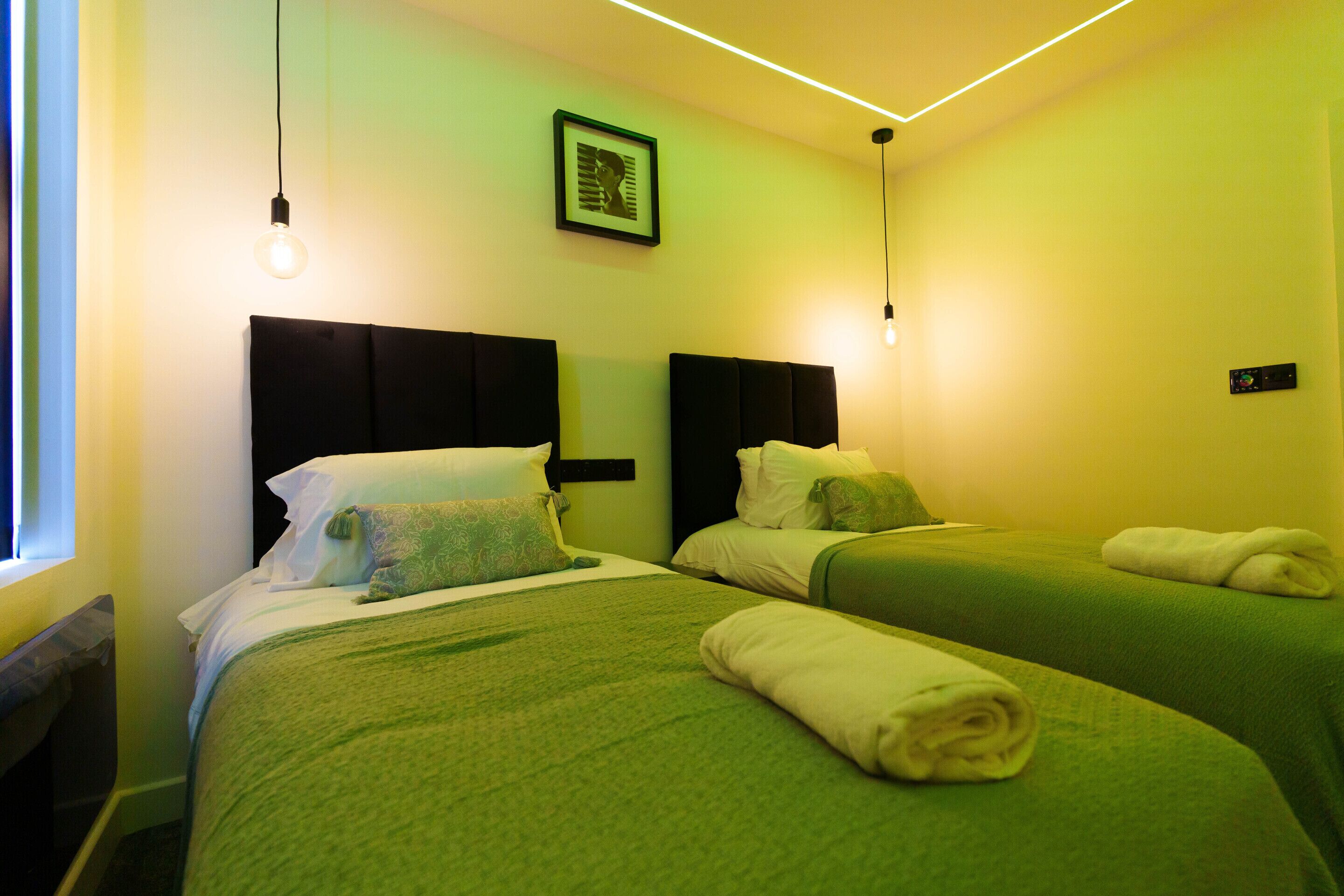 Ein Hotelzimmer mit zwei Einzelbetten, grüner Bettwäsche, einem gerahmten Bild an der Wand und einem Nachttisch mit Lampe.