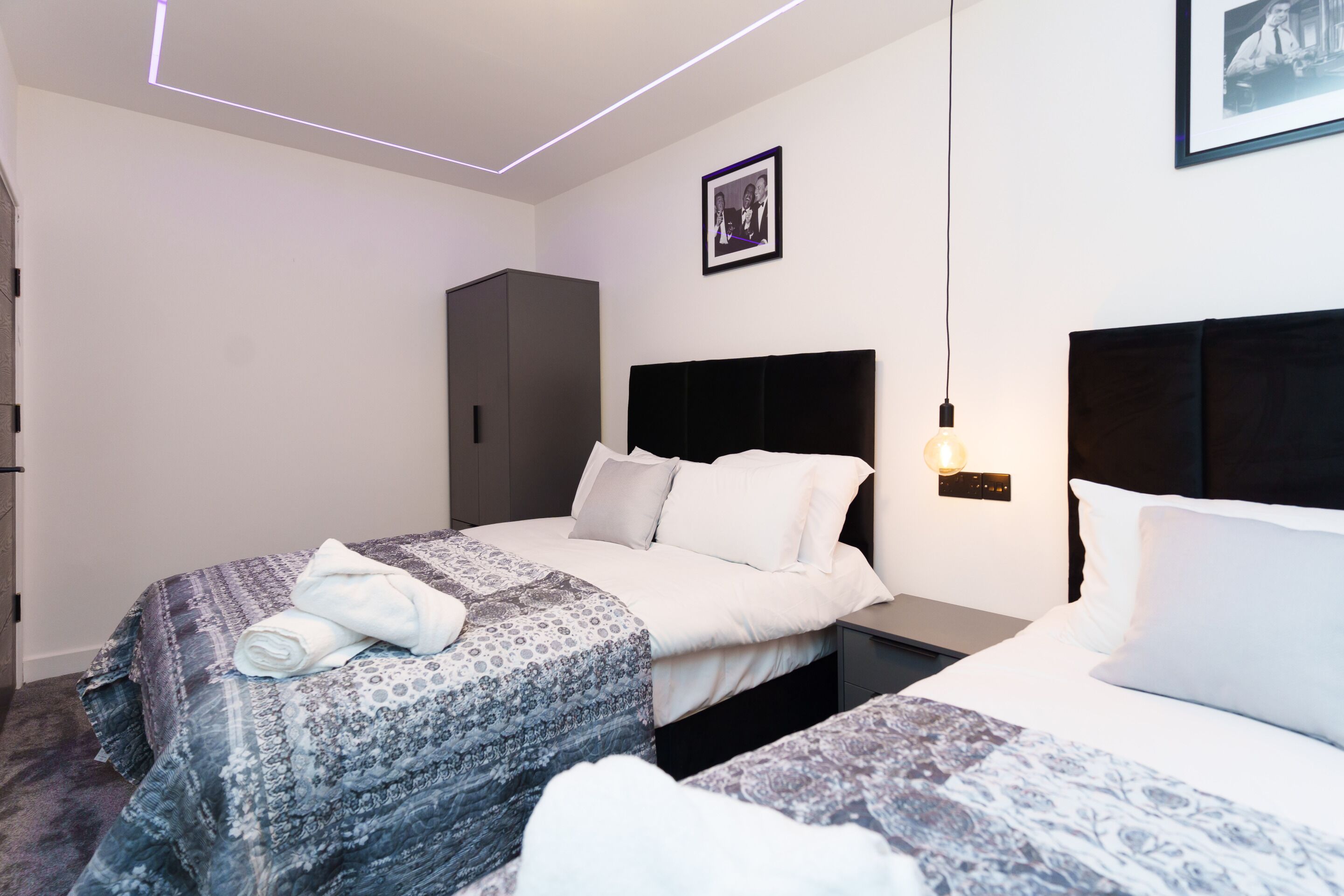 A hotel room with two beds, a wardrobe, a bedside table with a lamp, and a framed photo on the wall.