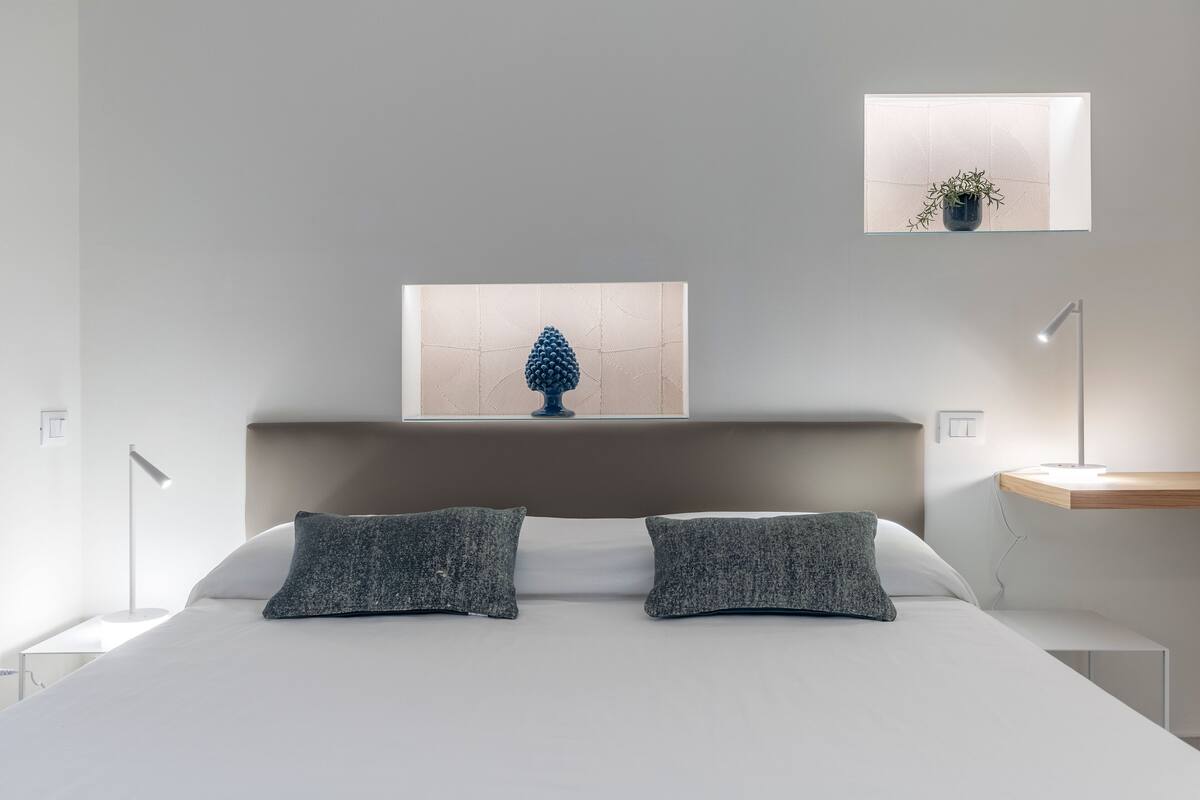 A modern bedroom with a large bed, two pillows, a bedside table, a nightstand, a wall-mounted shelf, and a window with a plant.
