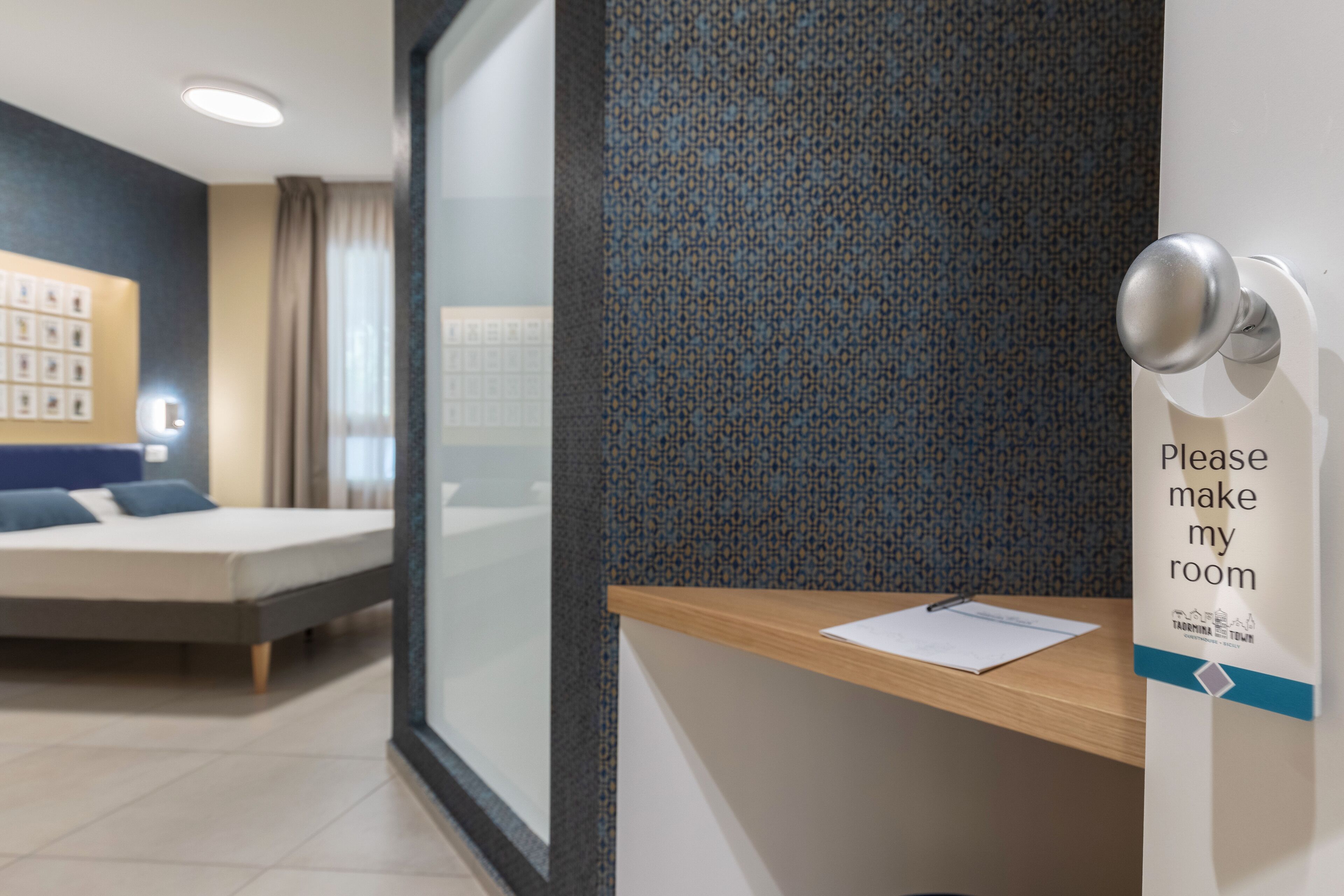 Une chambre d’hôtel avec un lit, un bureau sur lequel est posée une pancarte indiquant « Veuillez faire le ménage dans ma chambre », et un mur orné d’un motif.