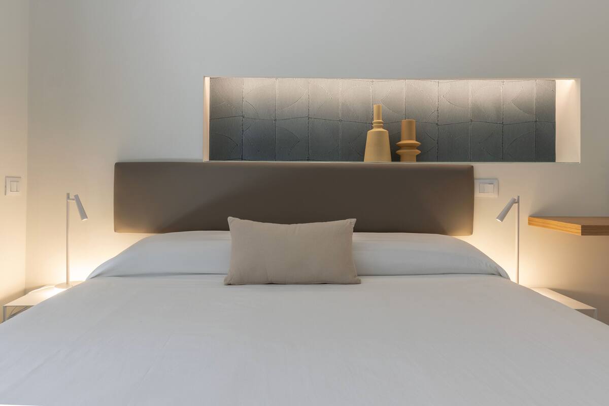 A neatly made bed with a pillow, a bedside table, and a wall-mounted light fixture.