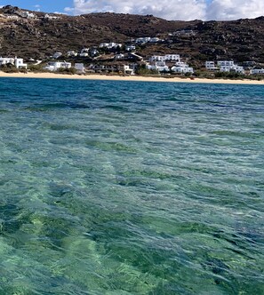 A coastal view with clear blue water, sandy beach, and white buildings on a hillside. - Villa Xanthos Située à 10 mn de Plaka Beach Dans un Cadre Paisible & Luxuriant (PLAKA)