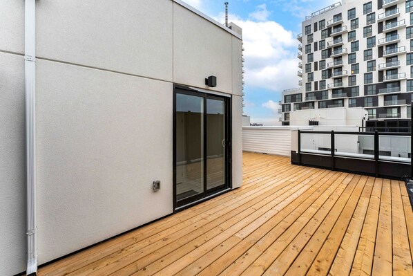 A rooftop terrace with a wooden deck, a glass door, and a modern building in the background.