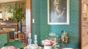 A dining area with a long table set for four, featuring teal patterned wallpaper, a framed portrait, and a bar cart with bottles.