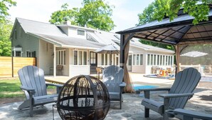 An outdoor patio with Adirondack chairs, a fire pit, and a covered area with a pool in the background.