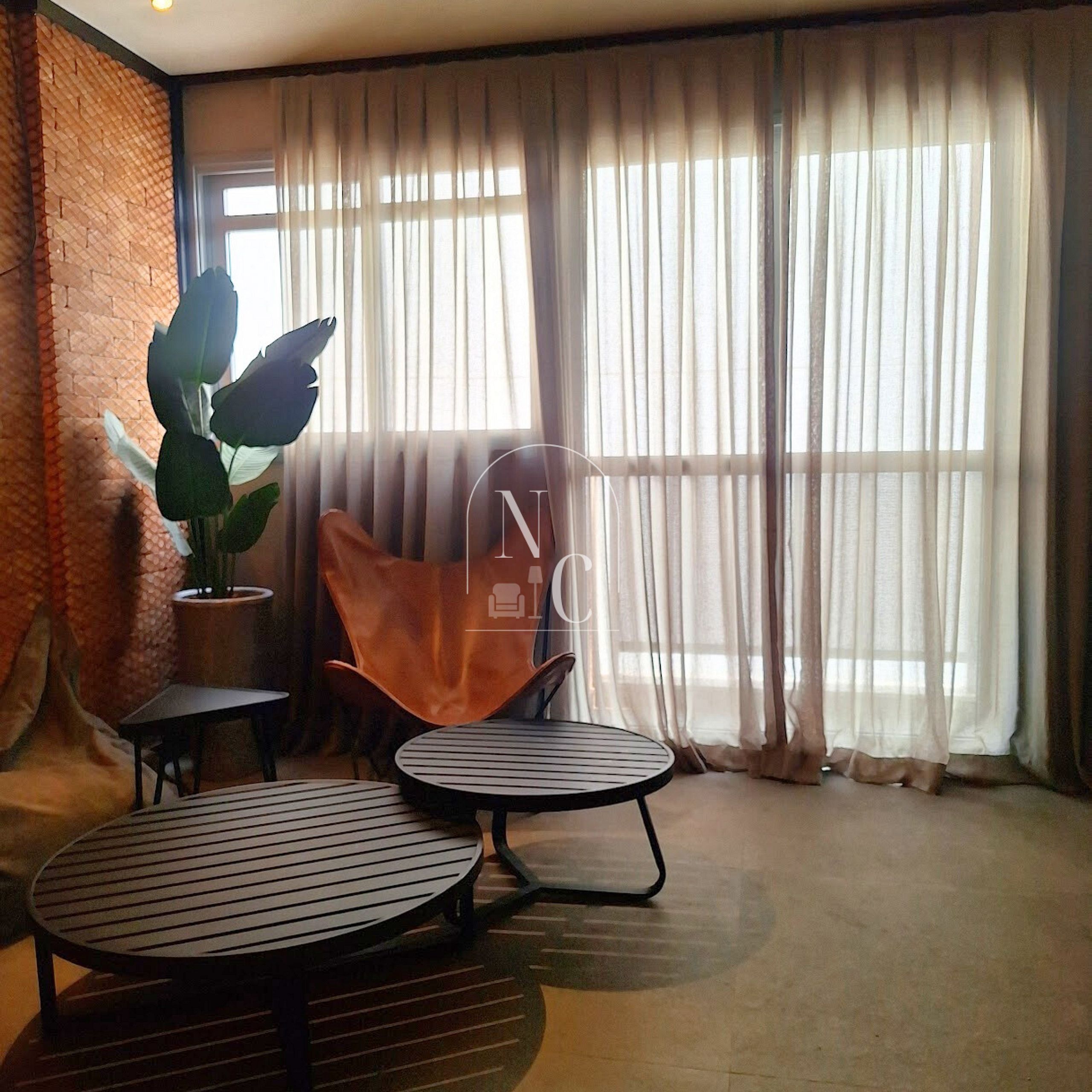 A room with a brick wall, a large window with sheer curtains, a potted plant, a leather chair, and two round tables.