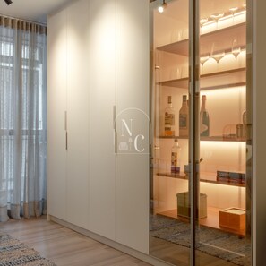Ein modernes Zimmer mit Kleiderschrank, einer Vitrine mit Flaschen und einem großen Fenster mit durchsichtigen Vorhängen.