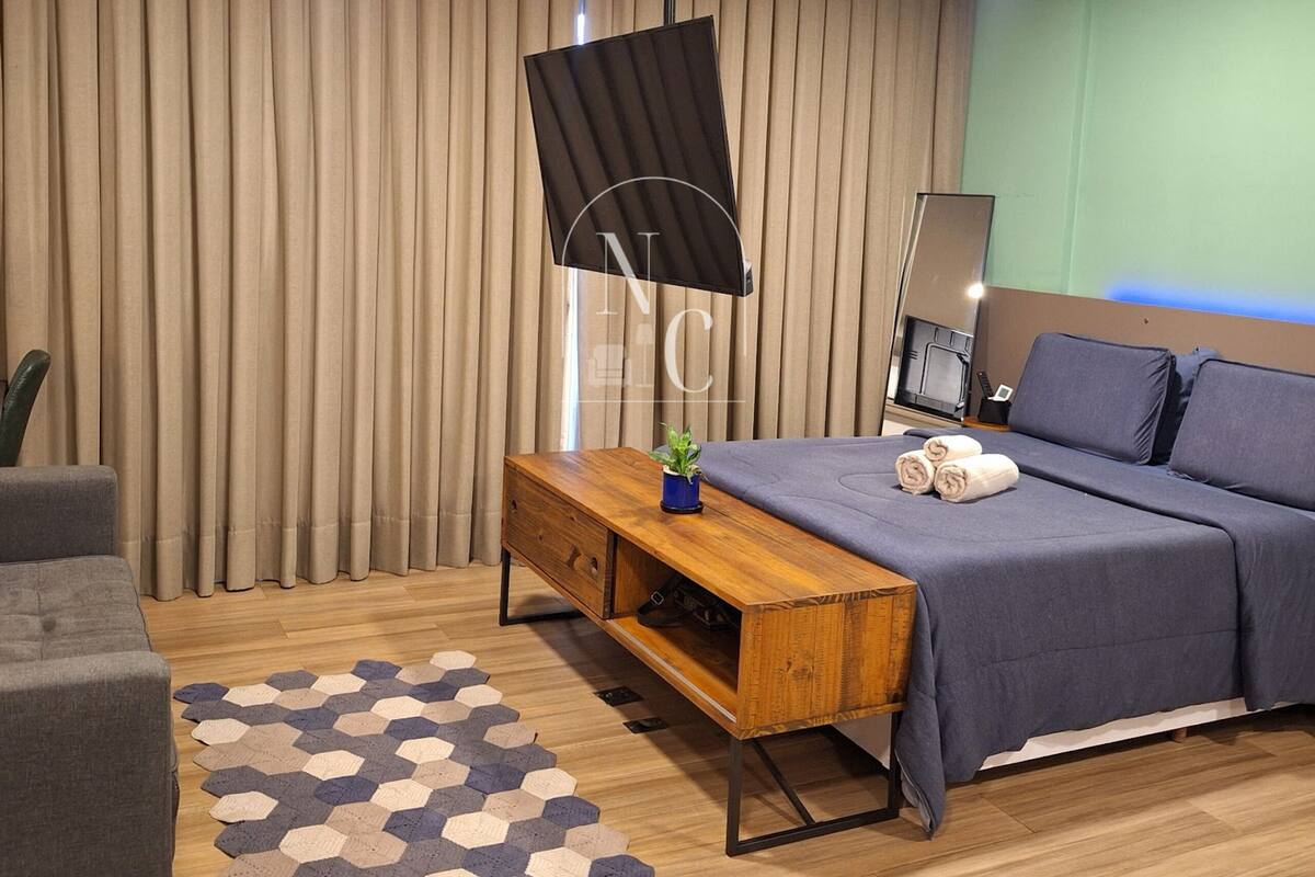 Quarto de hotel moderno com cama, um banco de madeira, escrivaninha, luminária e uma janela grande com cortinas.