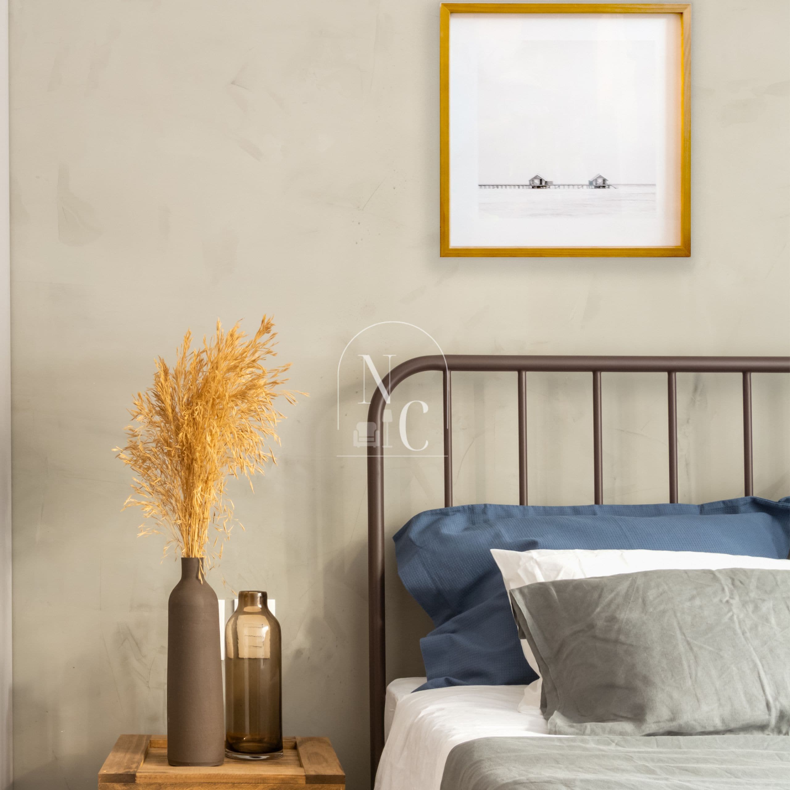 Een slaapkamer met een bed, een nachtkastje met een vaas en flessen, en een ingelijste foto aan de muur.
