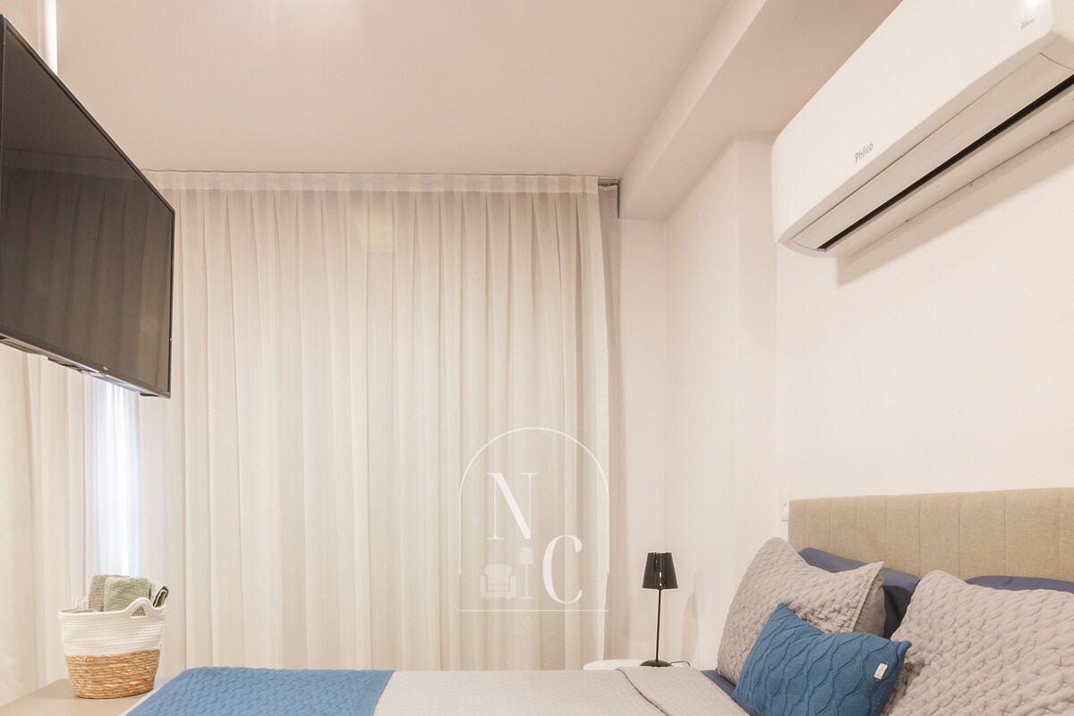 A bedroom with a bed, a bedside table, a lamp, a wall-mounted air conditioner, and a window with curtains.