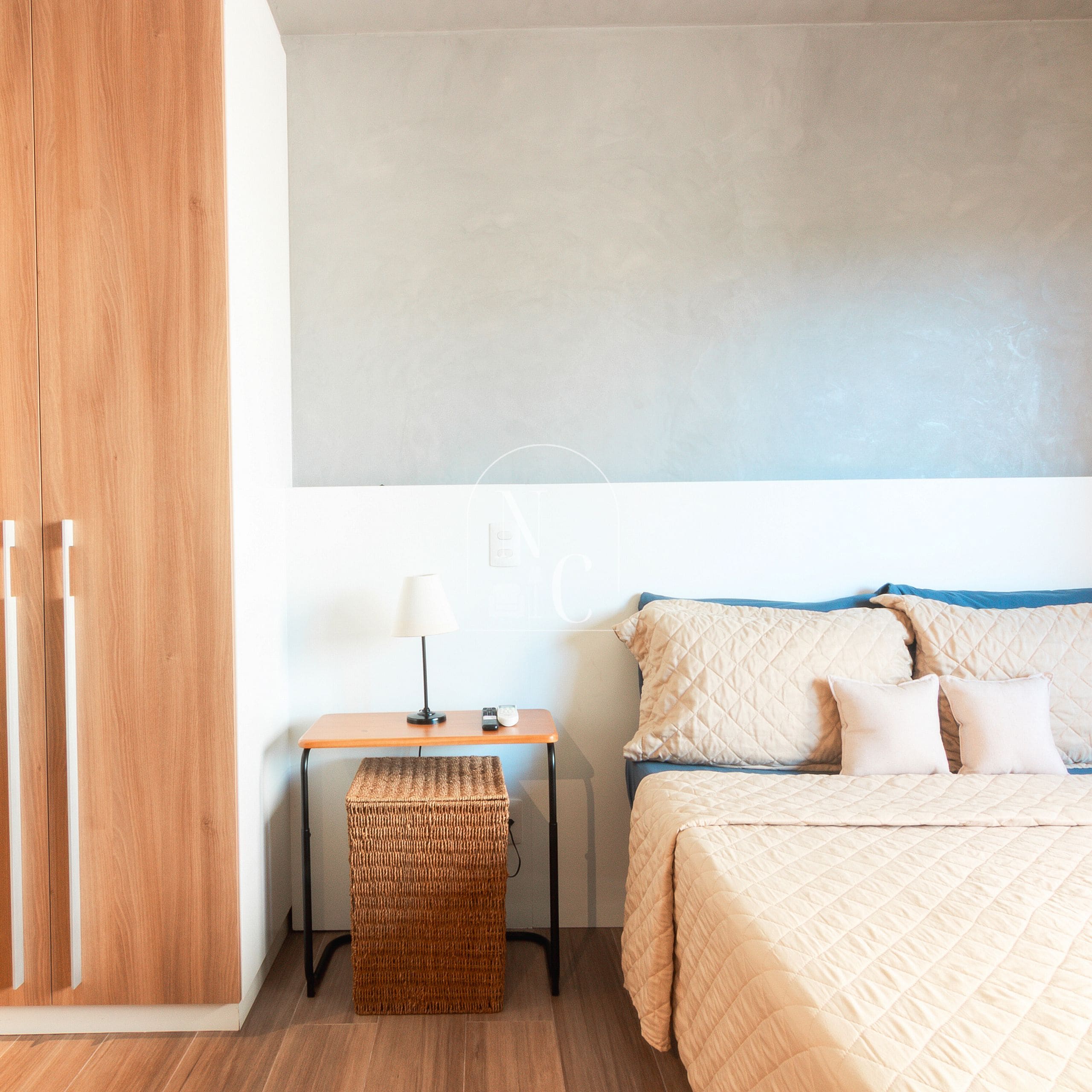 A bedroom with a bed, bedside table, lamp, and a small woven stool.