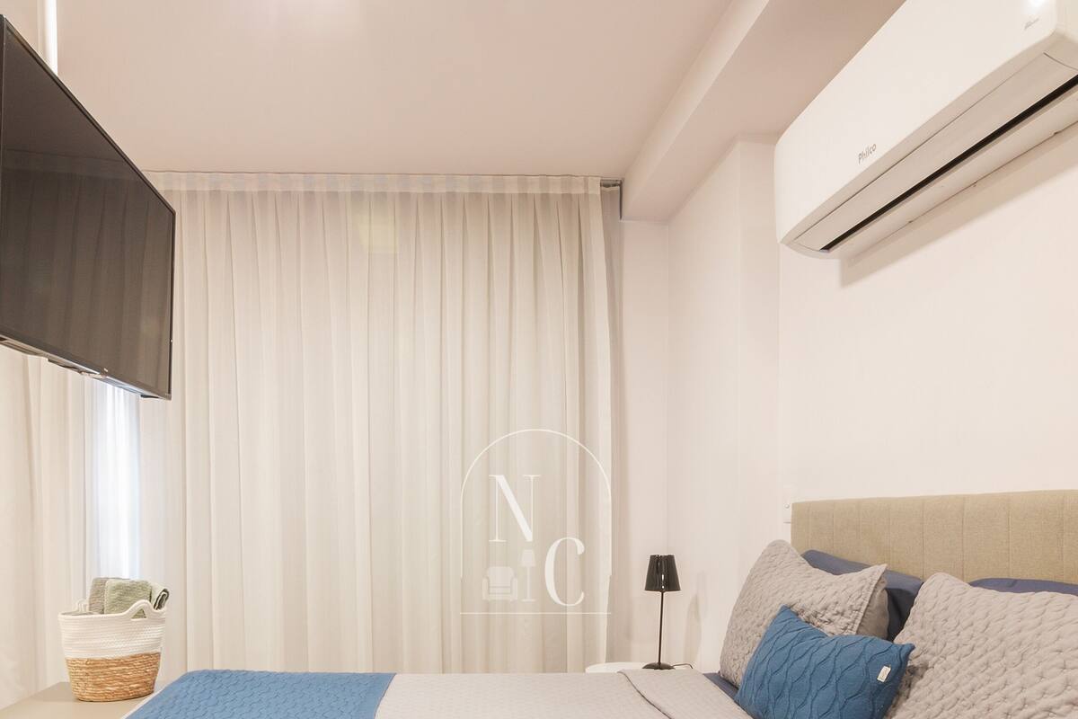 Quarto com cama, mesa de cabeceira, luminária, ar condicionado fixado na parede e janela com cortinas.