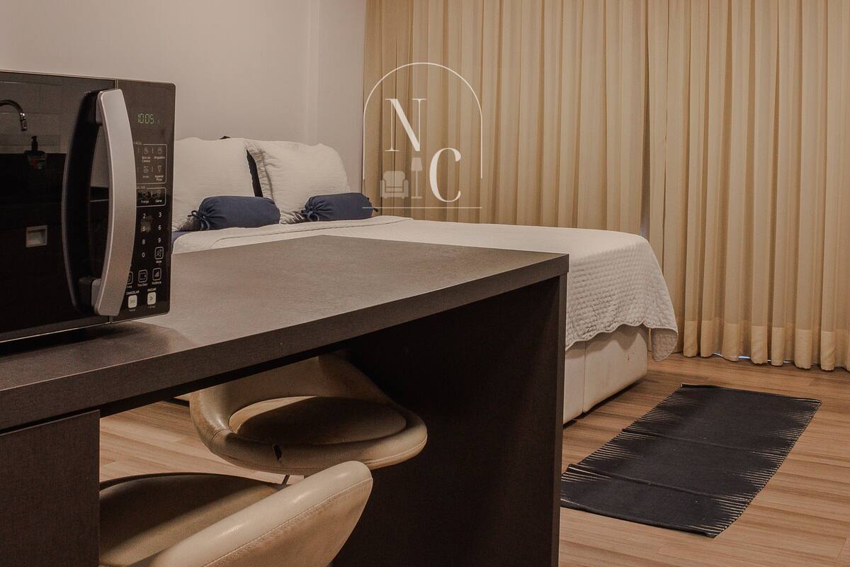 Quarto de hotel com cama, escrivaninha com micro-ondas, cadeira e janela grande com cortinas.