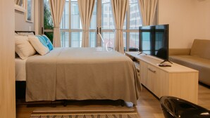 Habitació d'hotel moderna amb llit, televisió, sofà i grans finestres amb cortines.