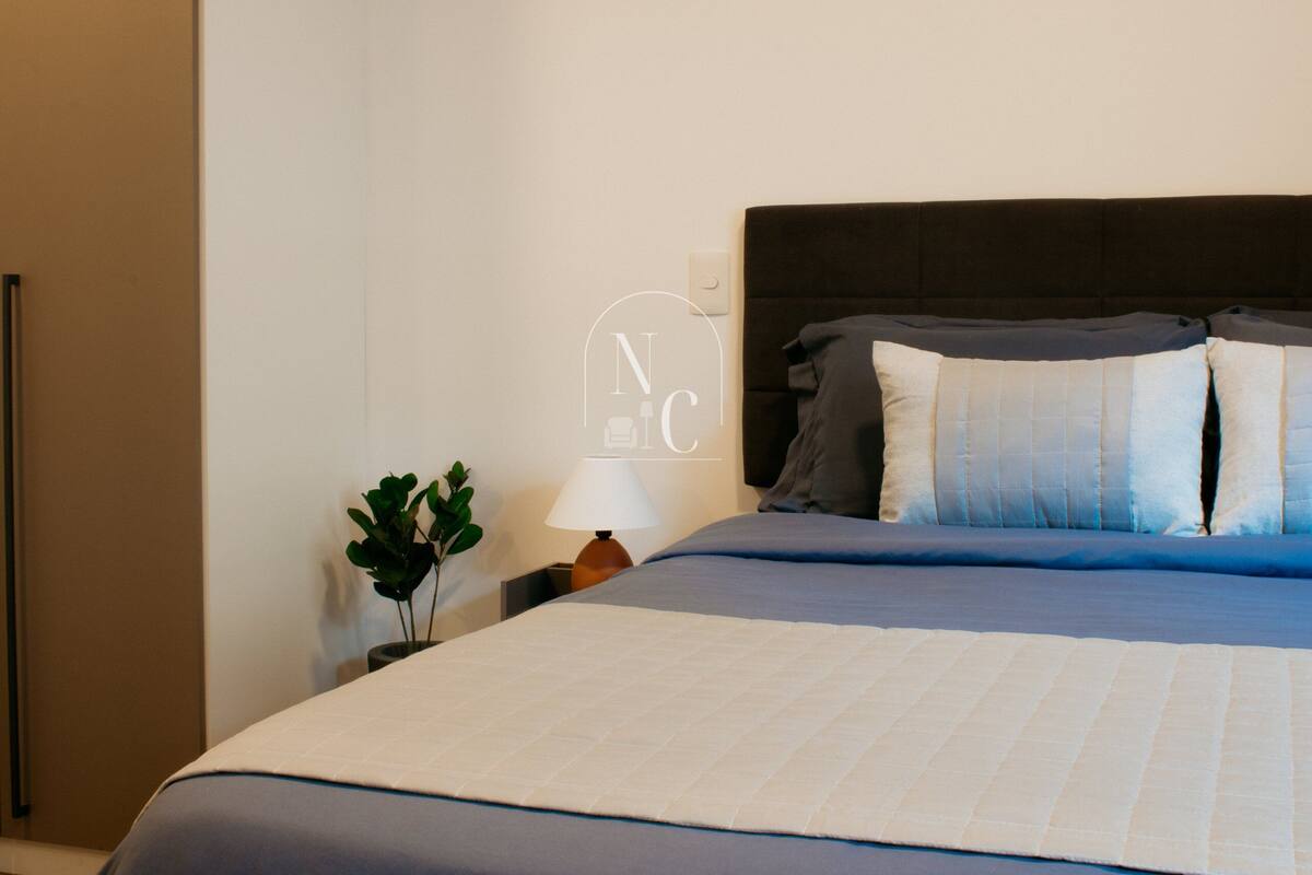 Uma cama bem arrumada com cabeceira escura, luminária de cabeceira e um pequeno vaso com planta.