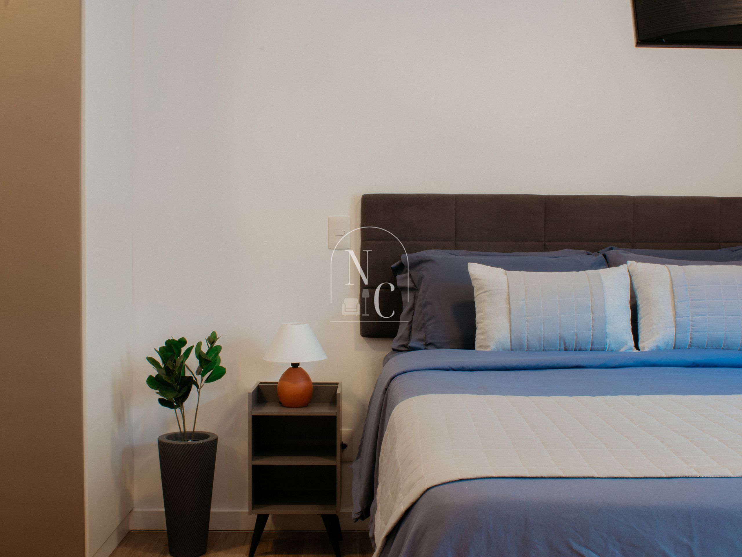 A bedroom with a bed, bedside table, lamp, and a potted plant.