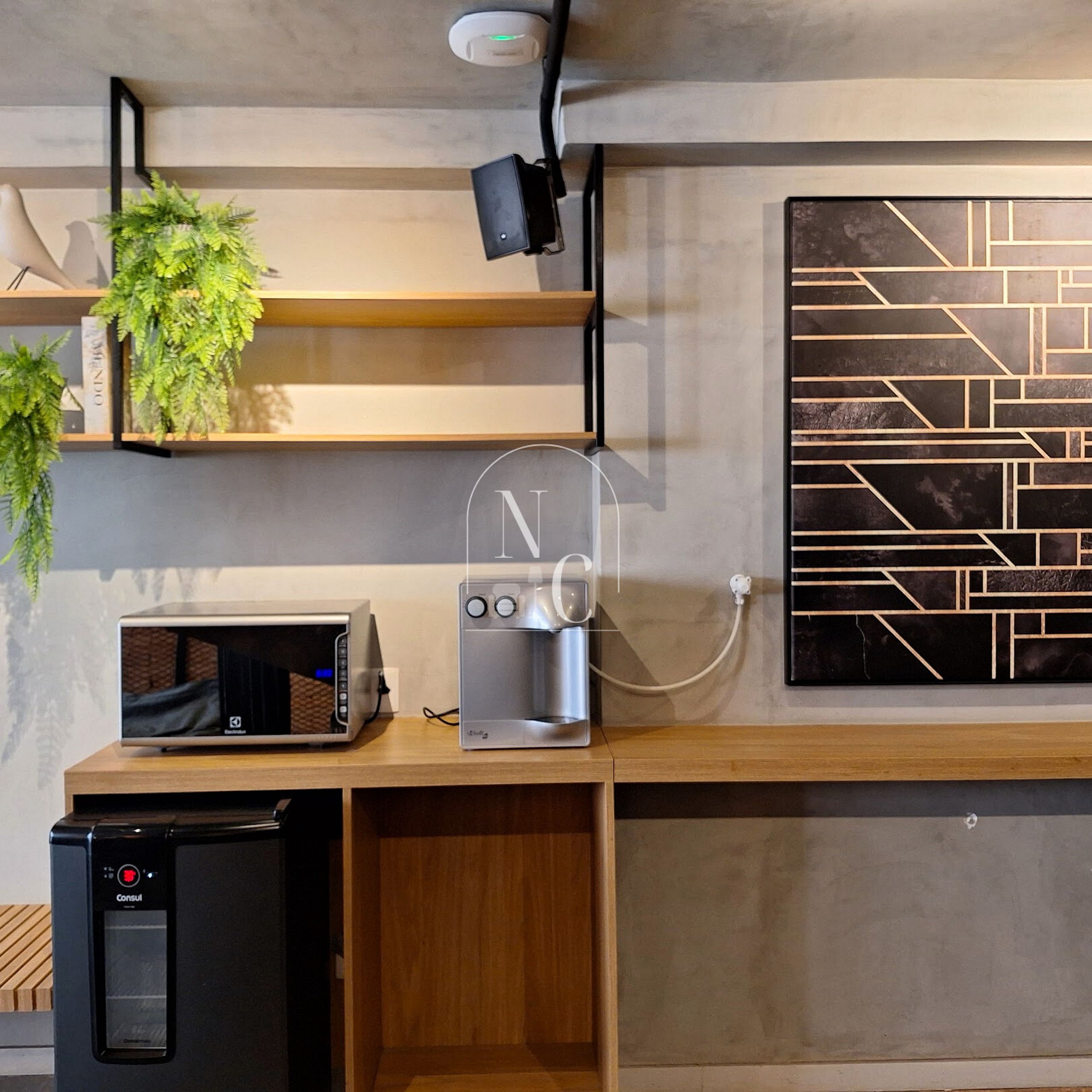 A kitchenette with a microwave, coffee maker, and a small refrigerator. There are shelves with plants and a wall-mounted light.